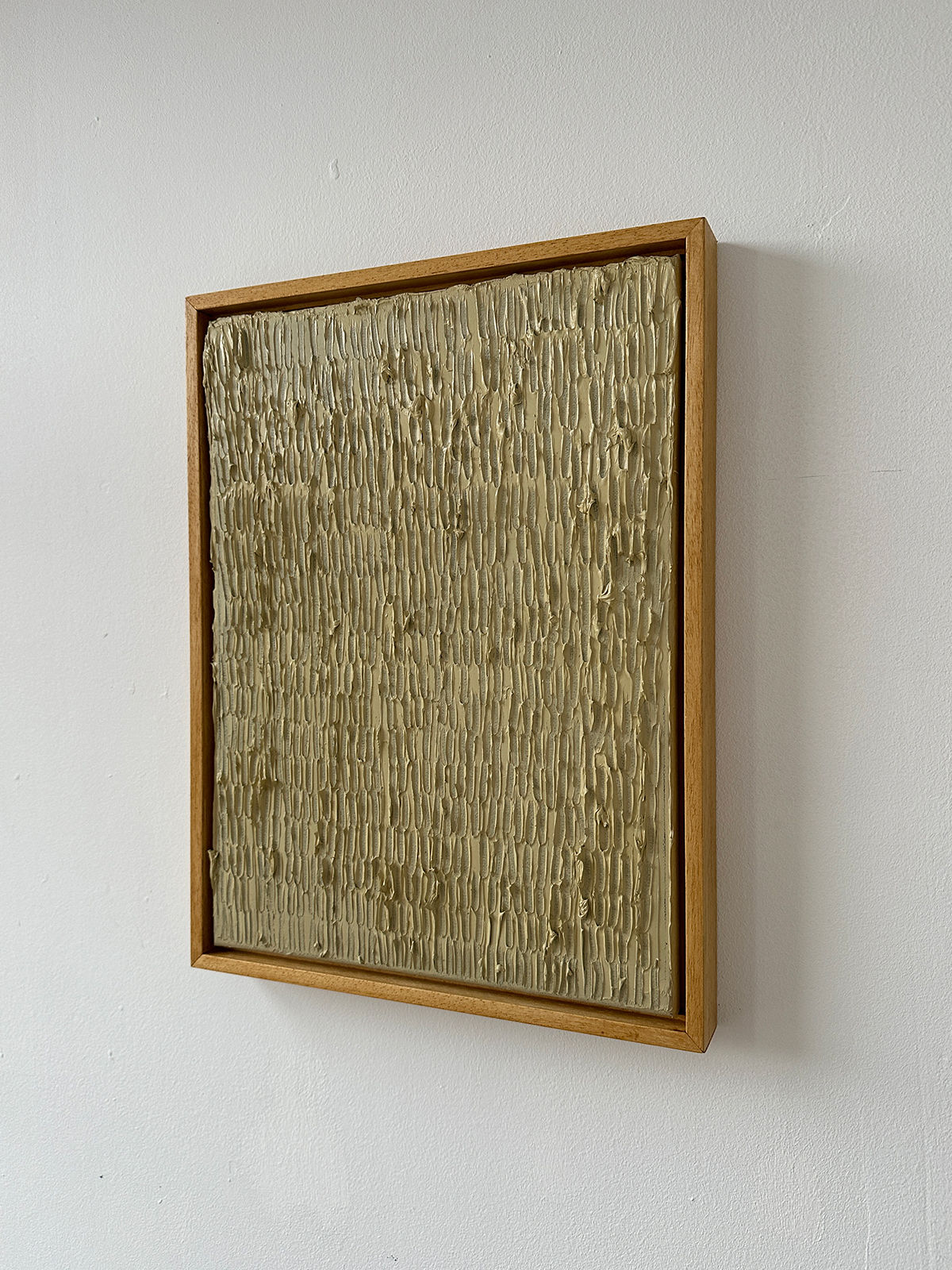 The "Buff Titanium" painting by Roi Elmaliah is a textured, square artwork showcasing vertical ridges on a beige surface. Crafted with oil on canvas, it is surrounded by a thin wooden border and mounted on a white wall.