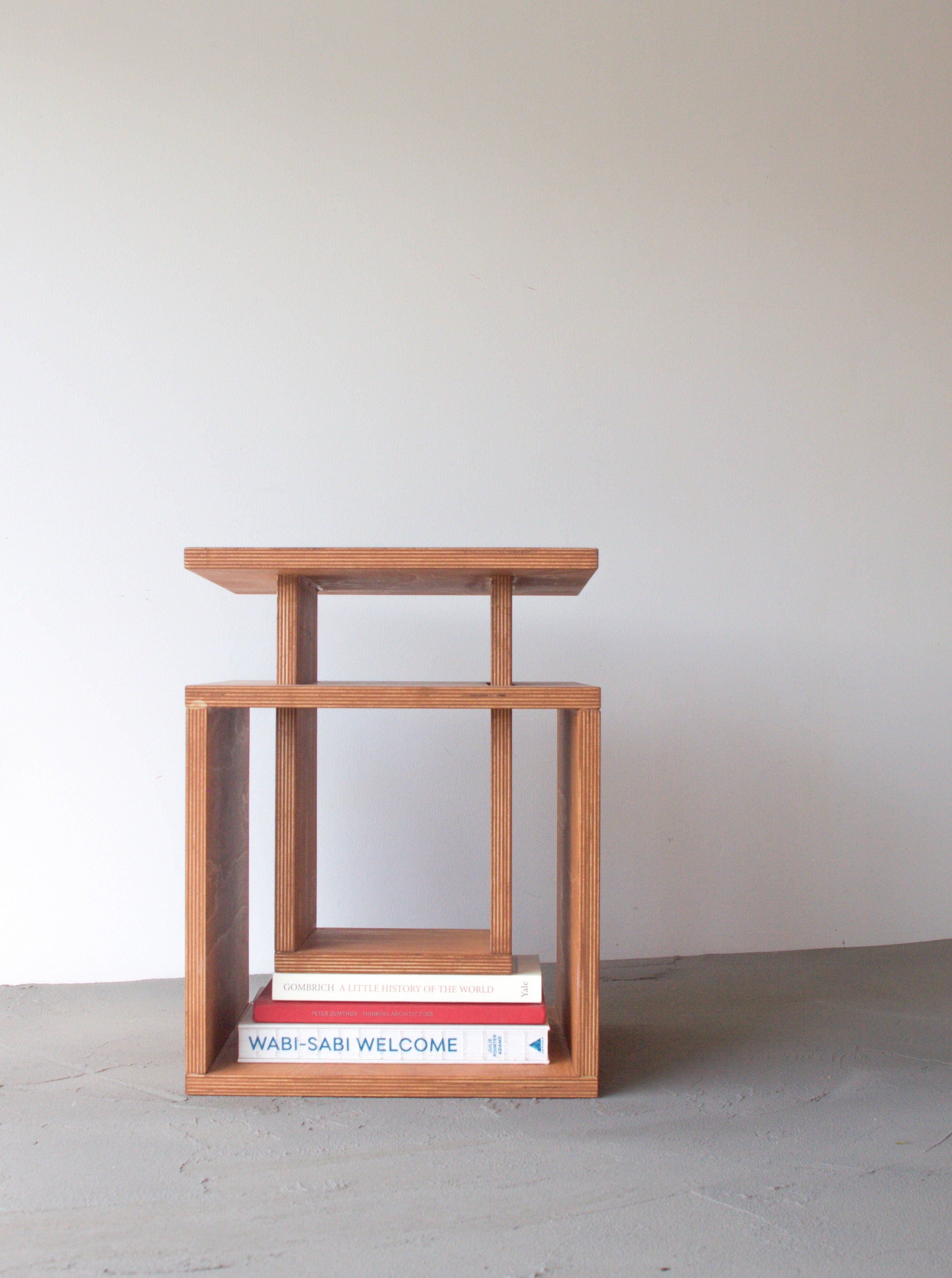 A minimalist Cubo side table by Edoardo Lietti Studio with a clean design, featuring a square base and a top platform, with a book titled "wabi-sabi welcome" placed on the lower shelf against a plain