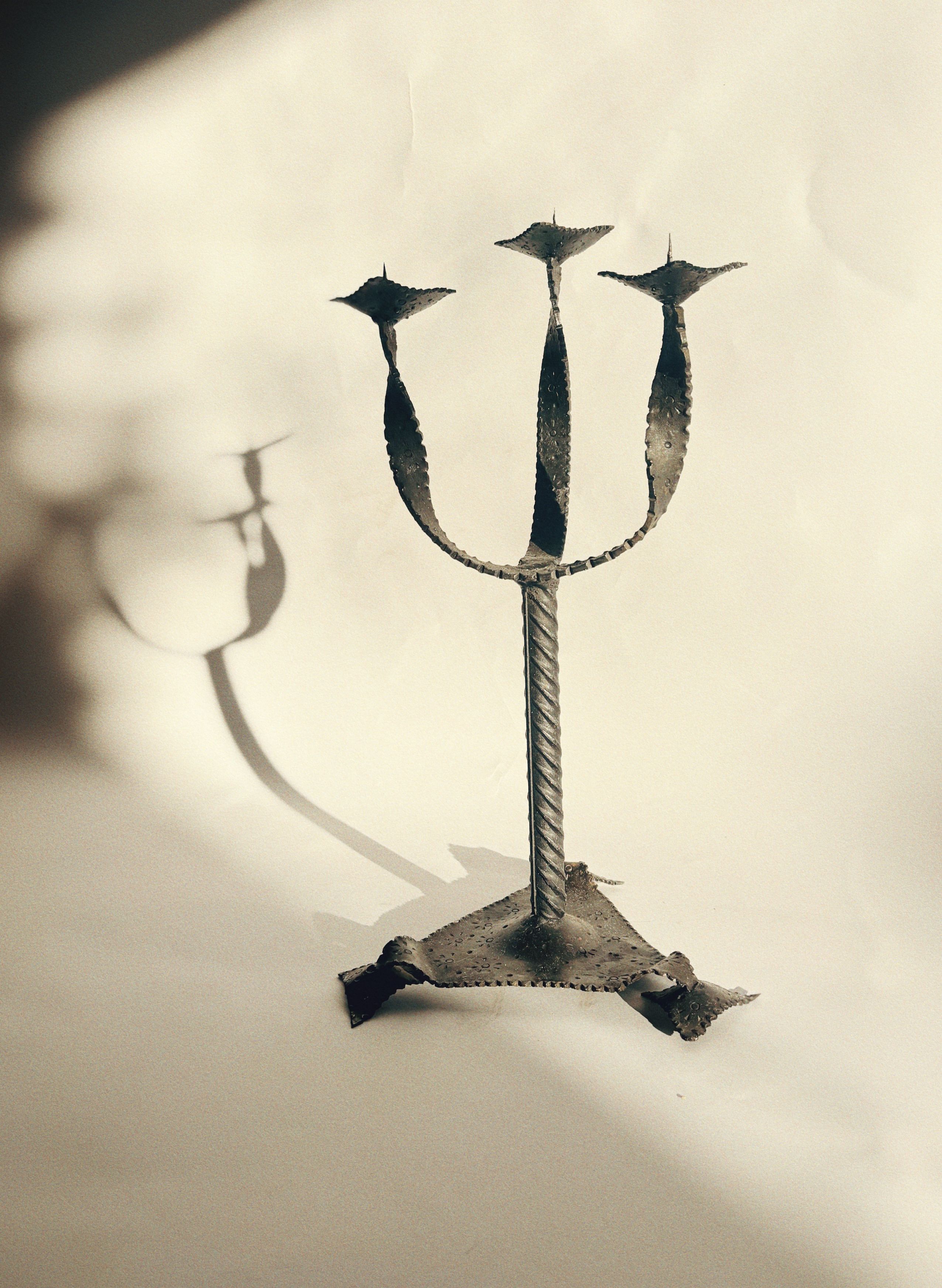 The Artisan Metal Candlestick by Huerto, designed in a rustic trident shape, evokes the feel of vintage functional artwork with its trio of prongs displayed against a simple backdrop. Crafted from hand-forged iron, this piece projects a clear shadow onto the surface below.