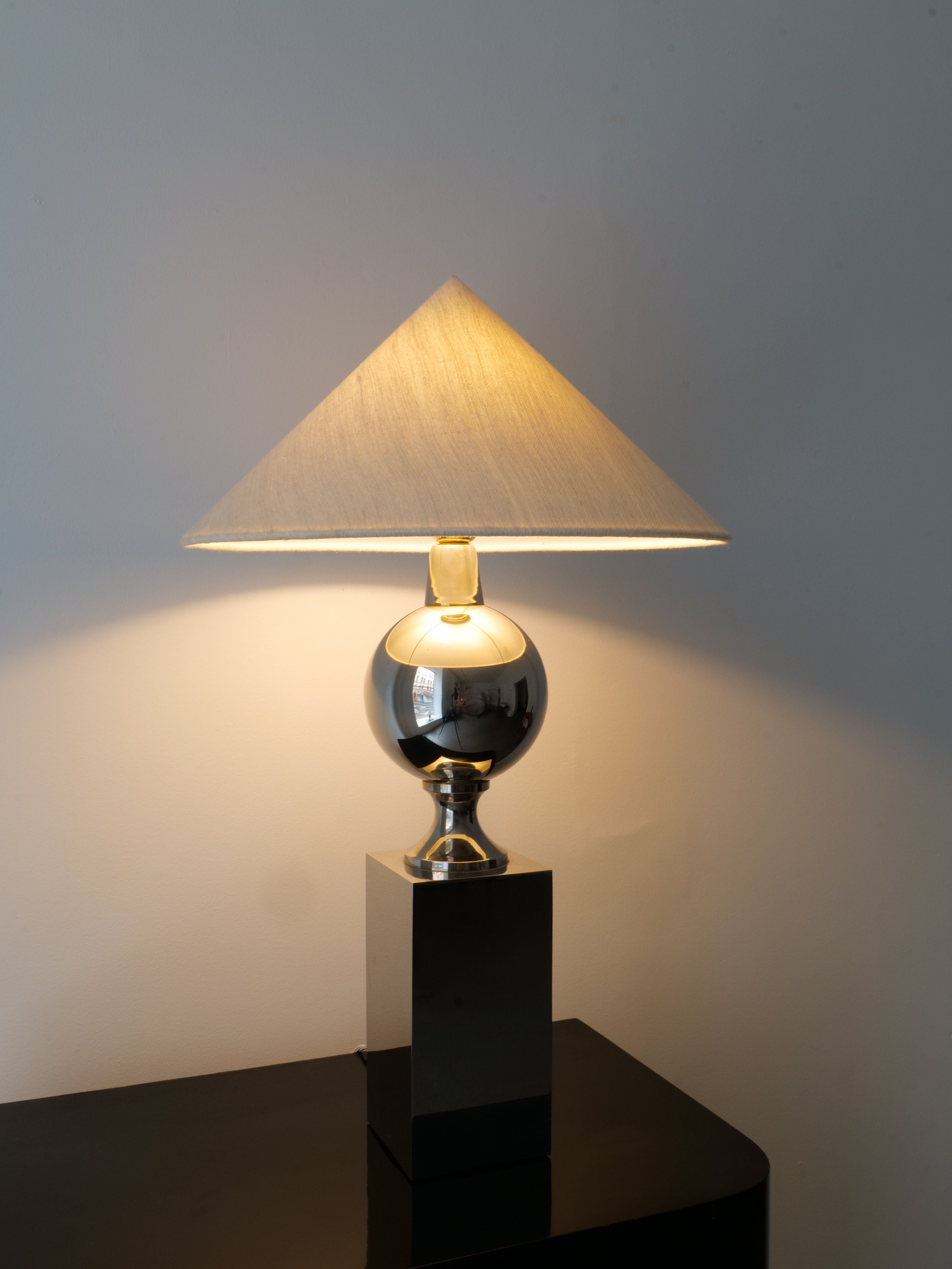 Introducing the Barbier Chrome Table Lamp by Collection Apart: This lamp features a chrome spherical base and a square pedestal, inspired by Philippe Barbier's sculptural designs for Maison Barbier. It is topped with a beige, cone-shaped shade that emits a warm glow on the wall behind, elegantly placed on a dark surface against a plain, light-colored background.