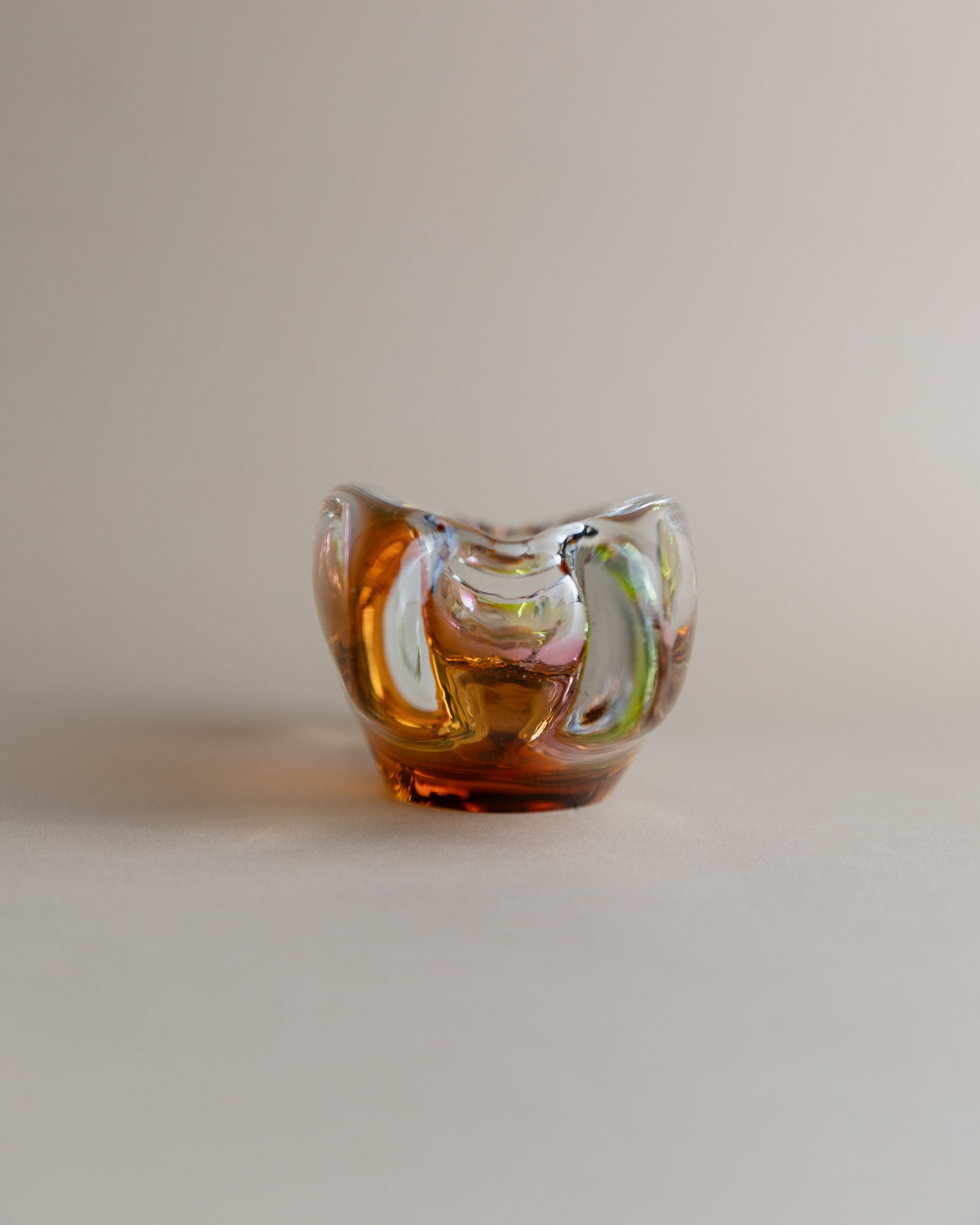 A small abstract glass ashtray by Josef Rozinek for Novy Bor, offered by Bottega Jacobs, features swirling amber, green, and blue colors on a neutral backdrop. It showcases a smooth, glossy surface with an organic shape typical of retro glassware artistry.