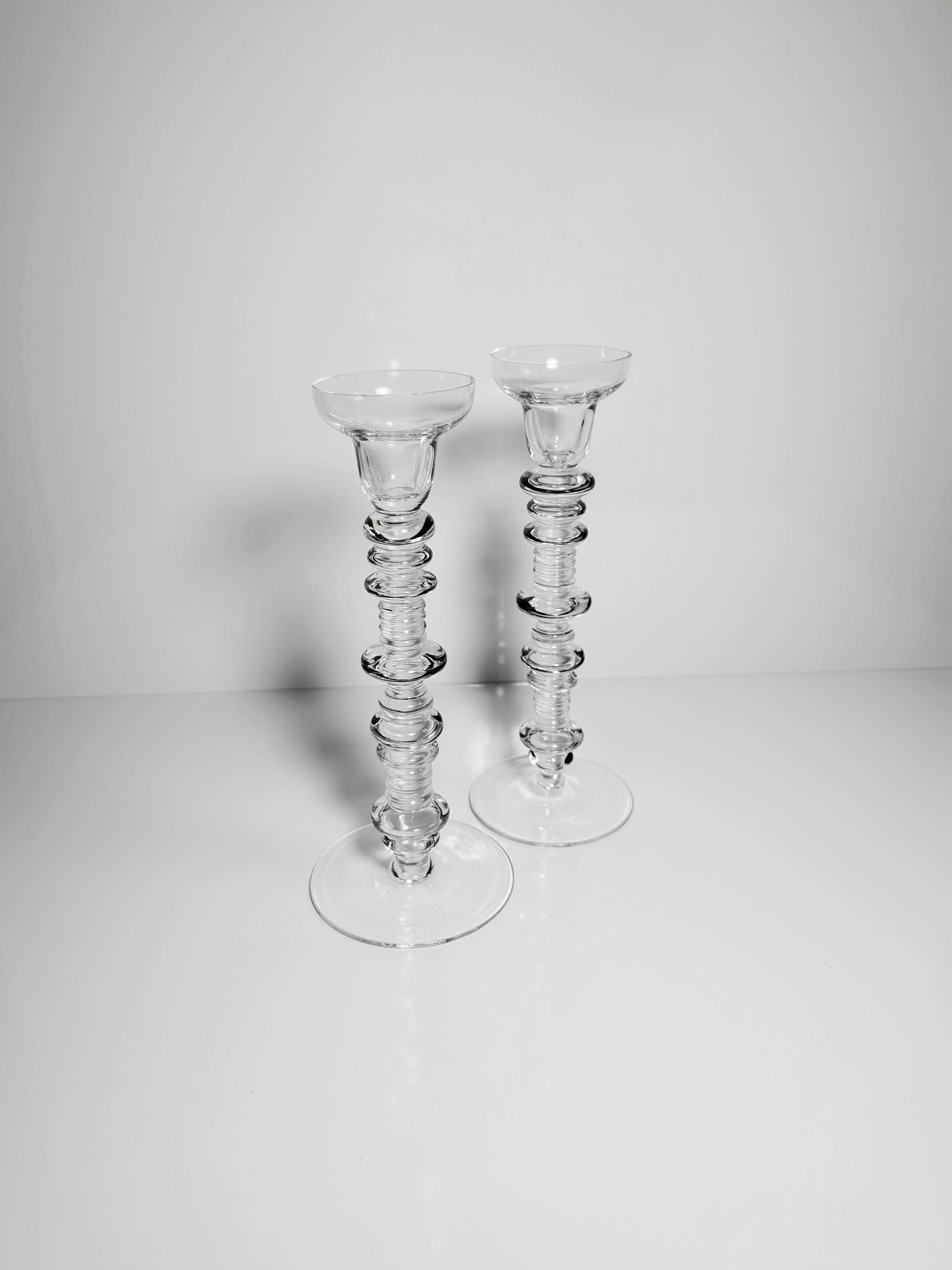 The Set of 2 Glass Candleholders by Peill & Putzler, available from Dodo Vintage, boasts a modern stacked ring motif on an elegant clear glass surface. These candlestick holders evoke a mid-century design, beautifully set against a smooth, pale gray background.