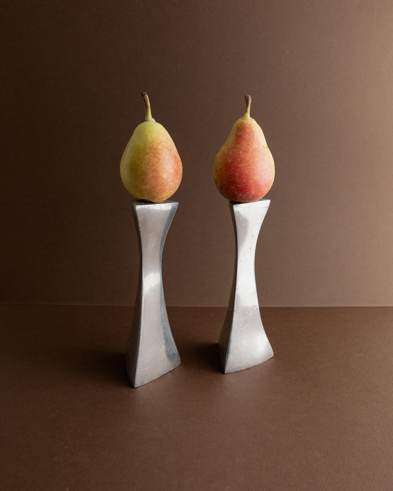 Two ripe pears are elegantly perched on the 2 Aluminium Candleholders 80s by Bottega Jacobs, featuring tall, slender metallic bases with a modern flair that evoke a retro-inspired ambiance against a plain brown background.