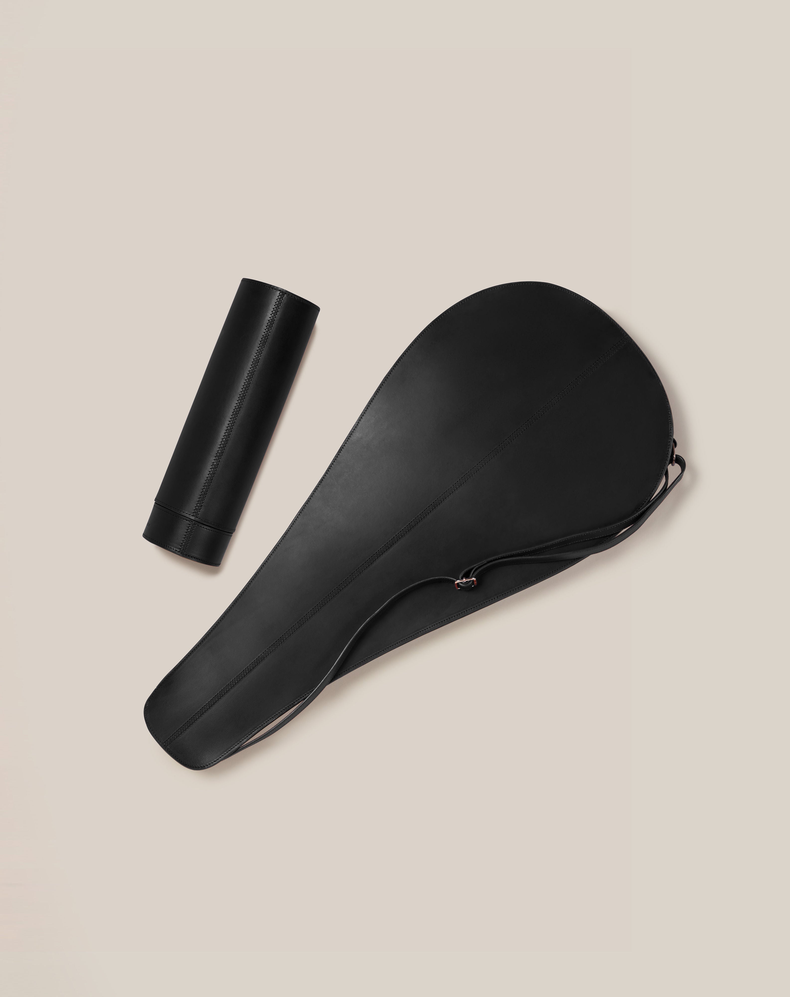 On a light beige background, a black leather cylinder case and shoehorn made from sustainable vegetable-tanned leather display bespoke British design by Paradise Row. The minimalist styled cylinder closes with a cap, while the shoehorn features a sleek, curved shape.