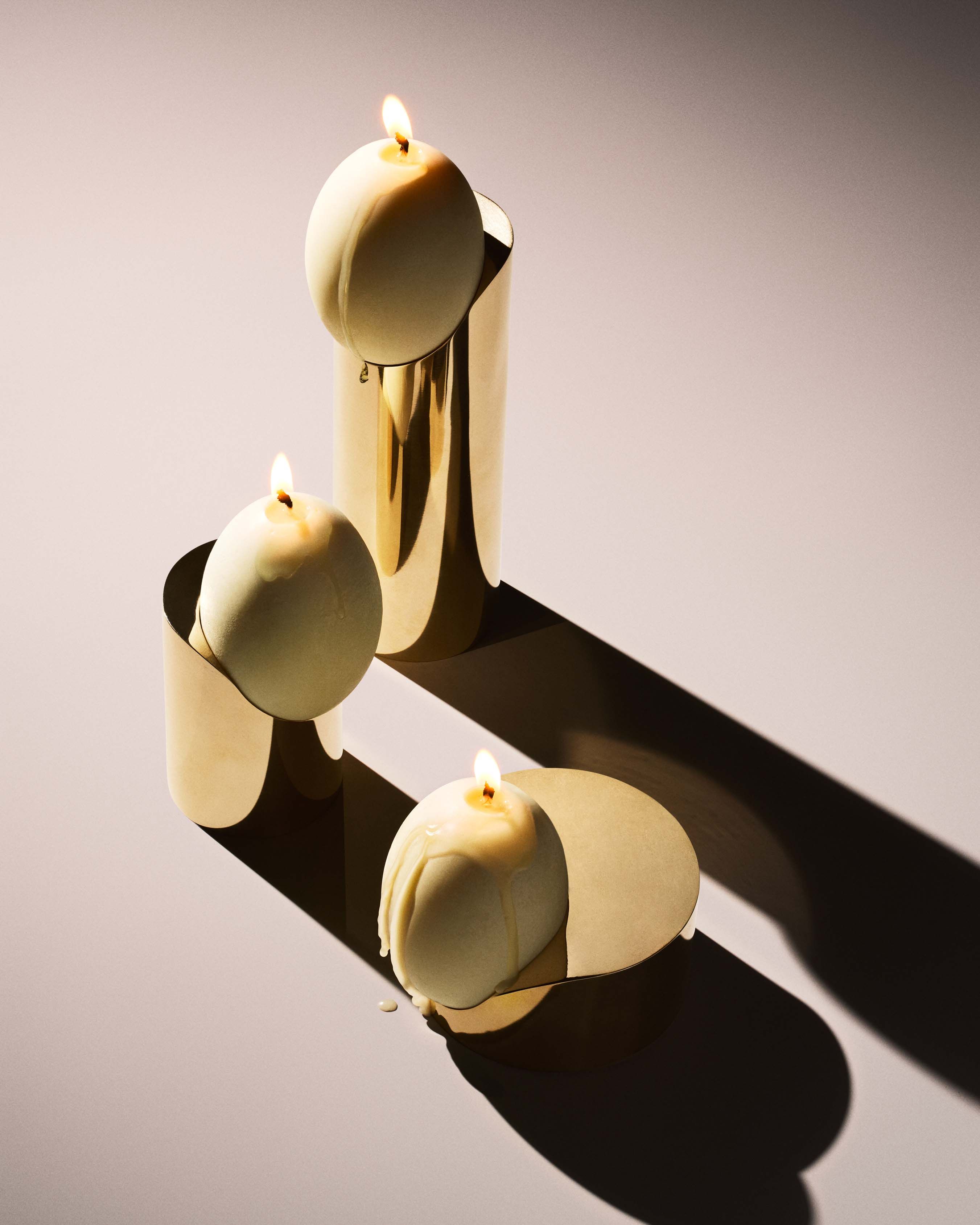 Three alight candles with dripping wax are placed on reflective, curved golden Low Perch Candle Holders from Tête Studio in a minimalist setting. The arrangement casts dramatic shadows on the smooth surface, reminiscent of Noguchi sculptures, creating a modern and elegant visual effect.