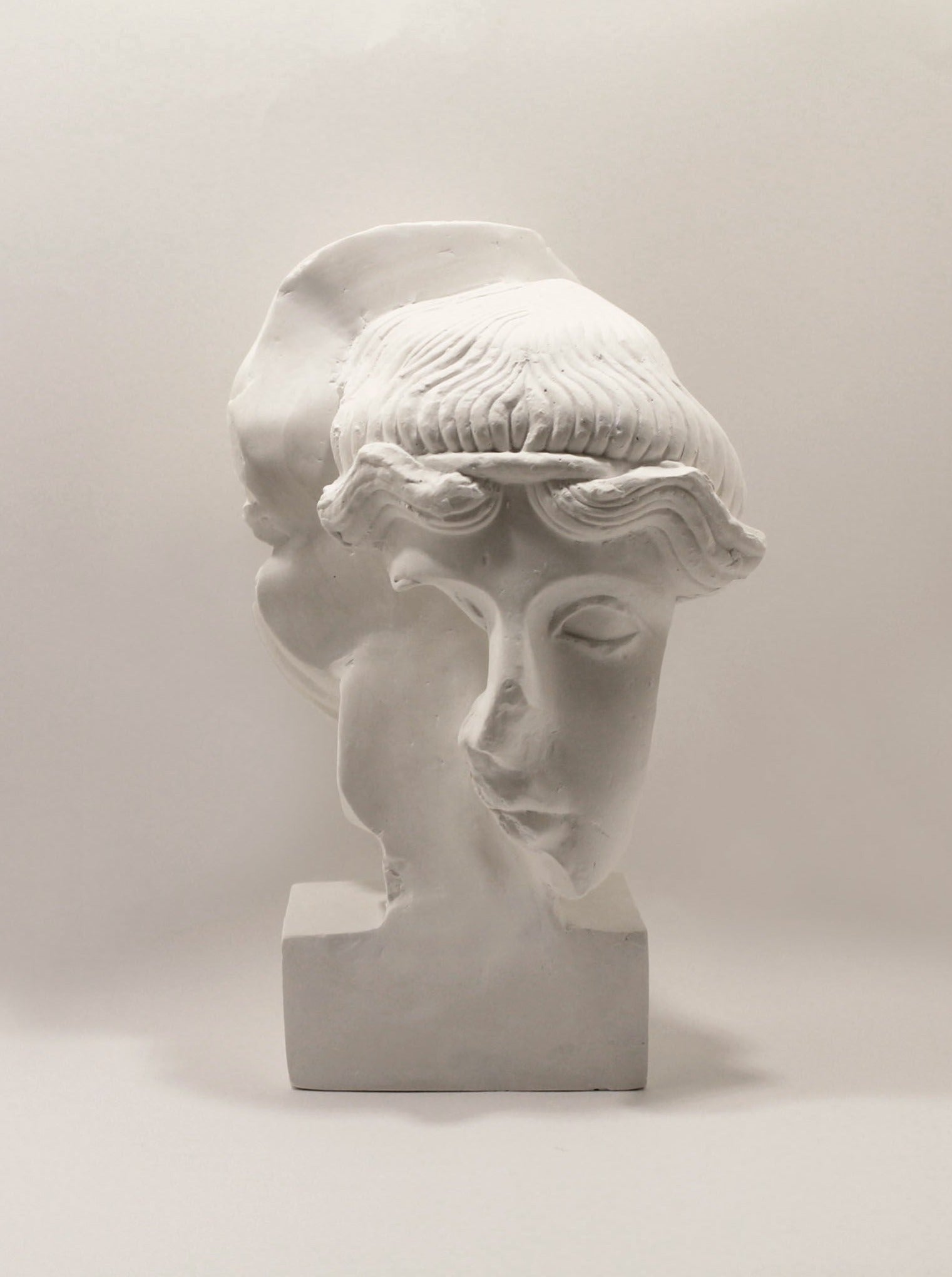 The Fragment #9 Sculpture 2021 by Marina Mankarios is a white plaster piece depicting the dual faces of a person, potentially symbolizing duality or two sides. One face gazes forward while the other is turned sideways, both displaying classical and serene expressions with intricately detailed hair and facial features.