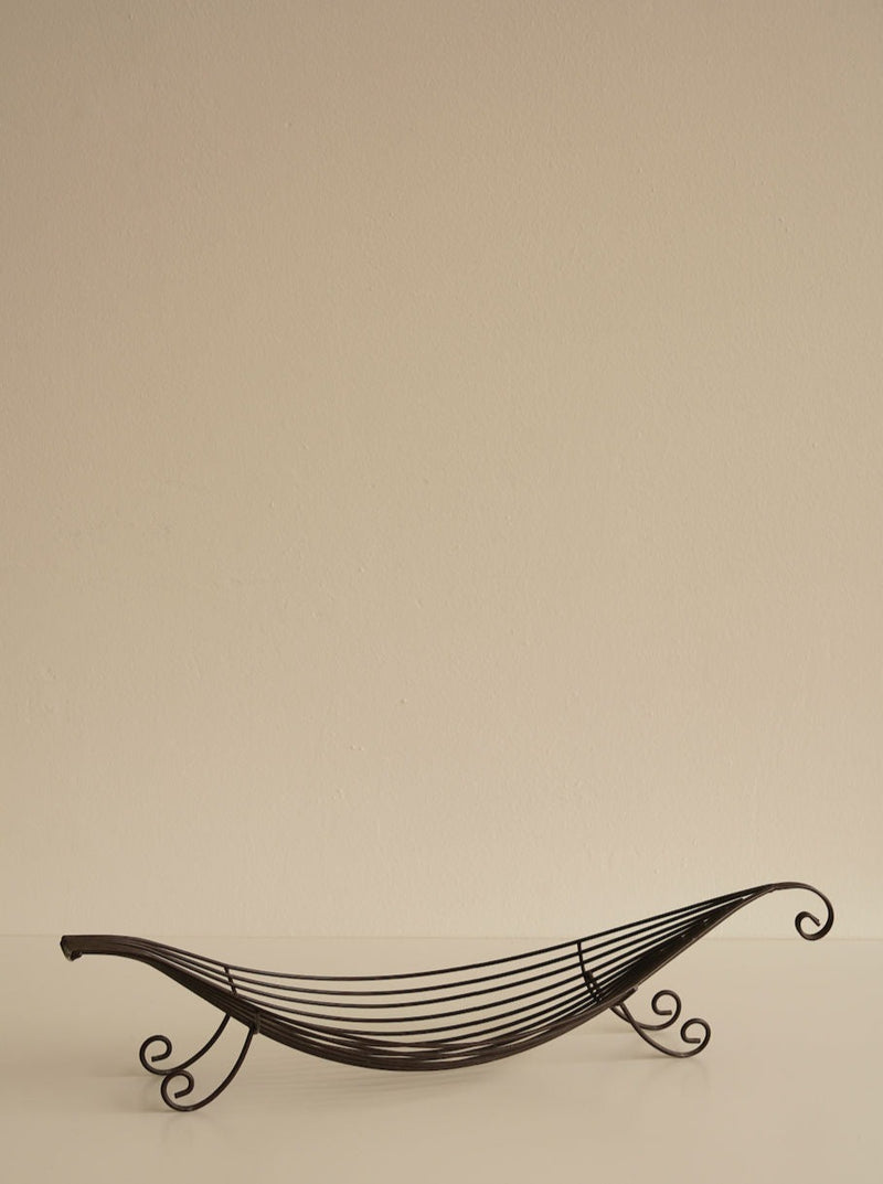 An Iron Swirl Fruit Bowl by Les Objets Amsterdam with an elegant, curved design sits empty against a plain beige background. The elegant fruit bowl features intricate, flowing loops and swirl accents on the ends and sides, creating a sleek and artistic look.