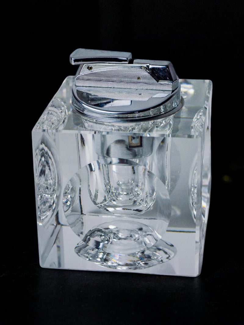 The Ice Cube Lighter by Antonio Imperatore 70s from Malamar Studio, featuring a clear cube-shaped design reminiscent of an ice cube with intricate circular engravings on each face, sits on a dark surface. The metallic top and mechanism create contrast against the transparent, reflective Murano glass-inspired look.