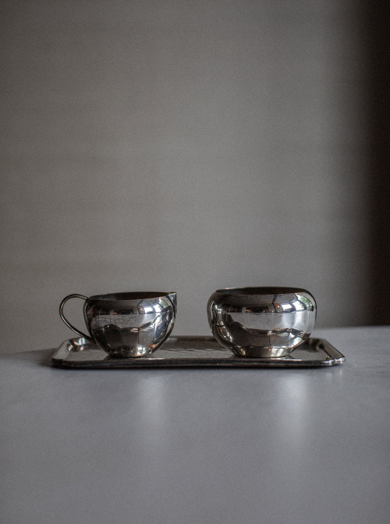 A minimalist photograph features an opulent Out For Lunch Art Deco Creamer & Sugar Bowl set placed on a matching rectangular tray. The background is softly blurred, allowing the focus to remain on the polished, reflective surfaces of the elegant tea set.