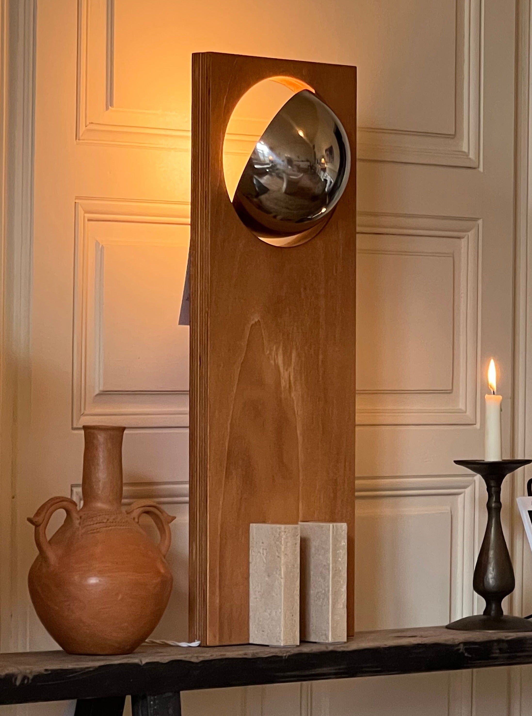 A Galileo 2.3 console displays a modern convex mirror mounted on a tall, wooden rectangular frame, accompanied by a rustic pottery vase and a lit table lamp, against a paneled wall. [Edoardo Lietti Studio]