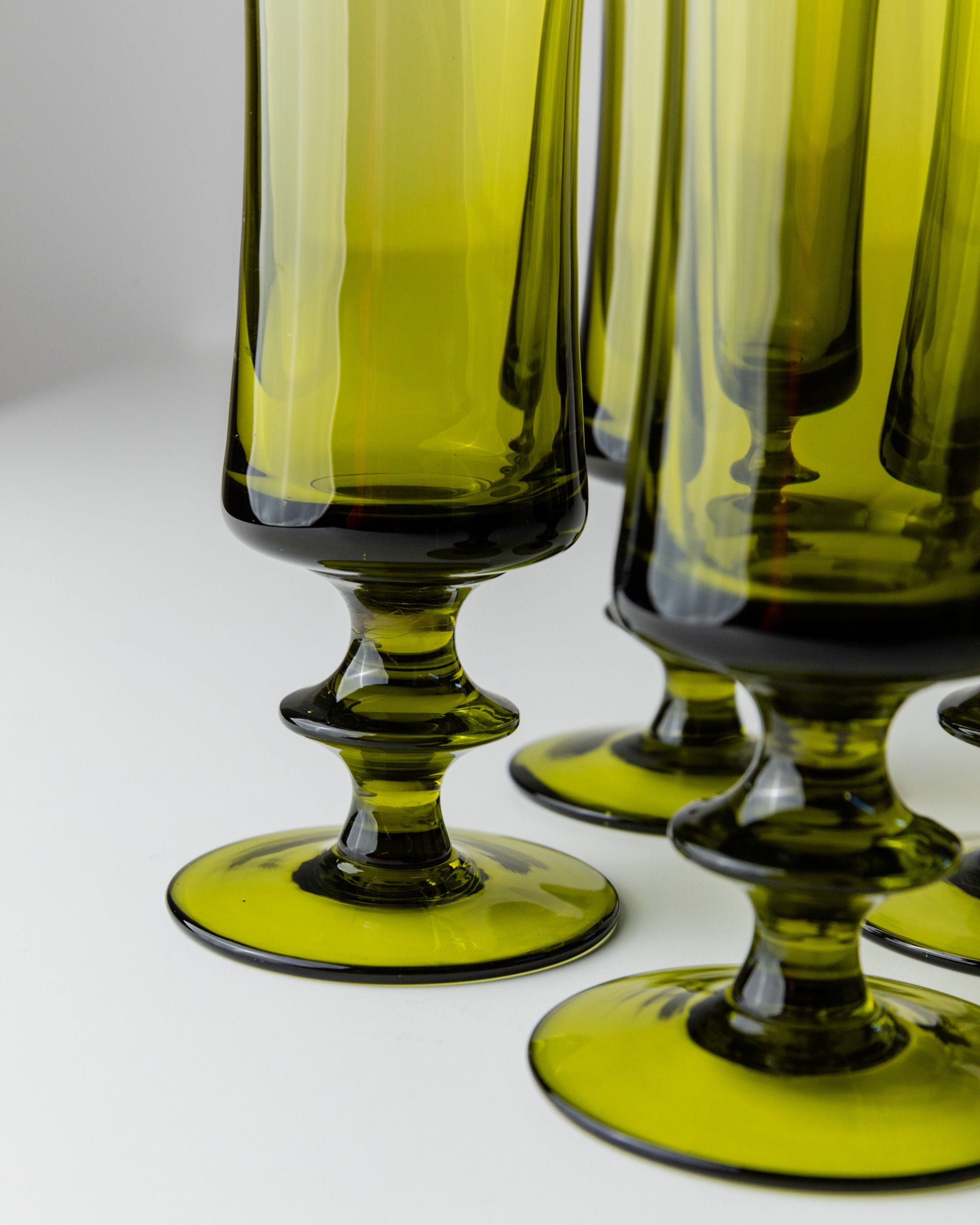 Close-up of Bottega Jacobs' 4 Murano Glasses in Green 70s with ornate bases on a white surface. The translucent glasses reflect light and evoke a retro chic vibe reminiscent of vintage Murano glass, creating a vivid and elegant appearance.