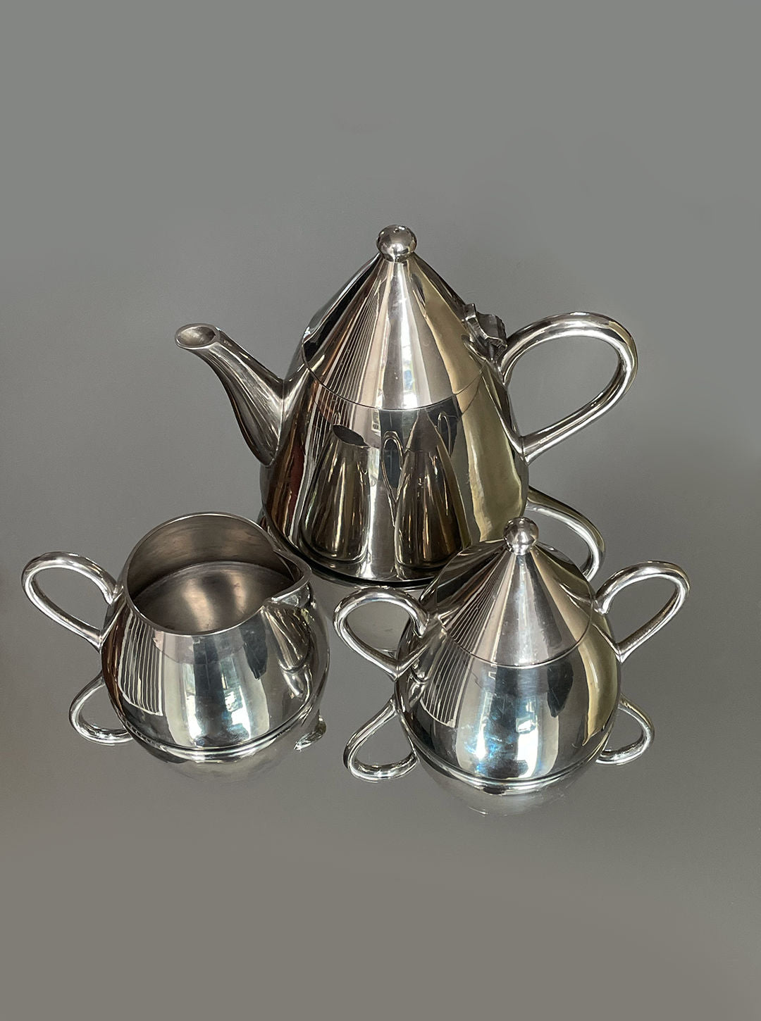 The Conic Modern Tea Set by Les Objoies features a shiny silver teapot, a lidded sugar bowl, and a lidless creamer. Each piece boasts smooth metallic finishes and curved handles, embodying contemporary design. Ideal for luxury tea rituals, the set stands out against a plain gray background.