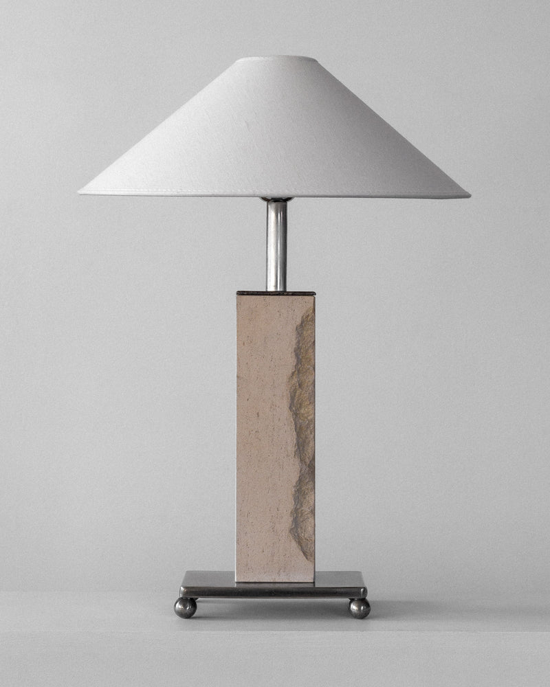 The room is graced by the Spigel Modernist Travertine and Metal Table Lamp, featuring a wide white conical shade. Its rectangular base has a stone-like finish with metallic accents, resting on a square platform with round feet. It exudes mid-century charm against a plain gray backdrop.