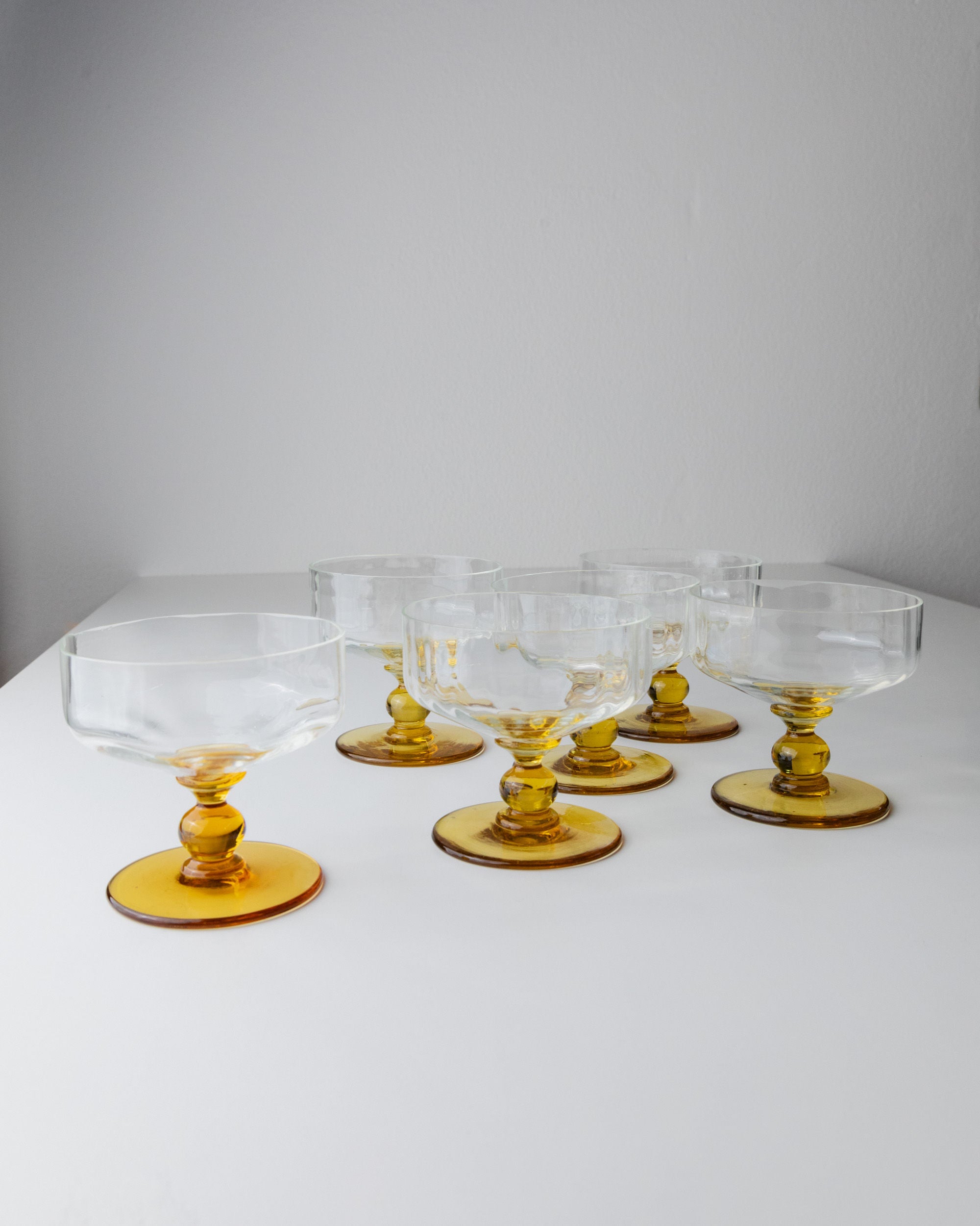 The Bottega Jacobs set of 6 Murano glass goblets from Italy's 70s era features clear glass with amber stems, arranged on a white surface against a light gray background, showcasing a simple and elegant design reminiscent of mid-century Italian style for a minimalist and contemporary look.