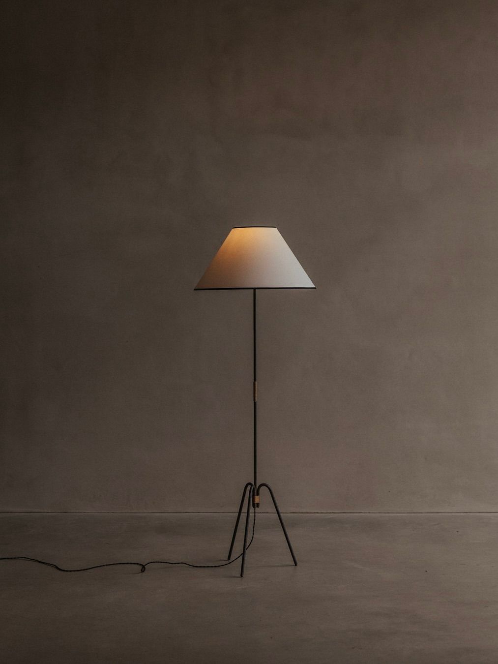 The Bicci de' Medici "Manner Of Royere & Adnet" Wrought Iron Tripod Lamp stands against a textured beige wall. This mid-20th century floor lamp with a white conical shade casts warm light from its slender black base. The cord is visible on the concrete floor.