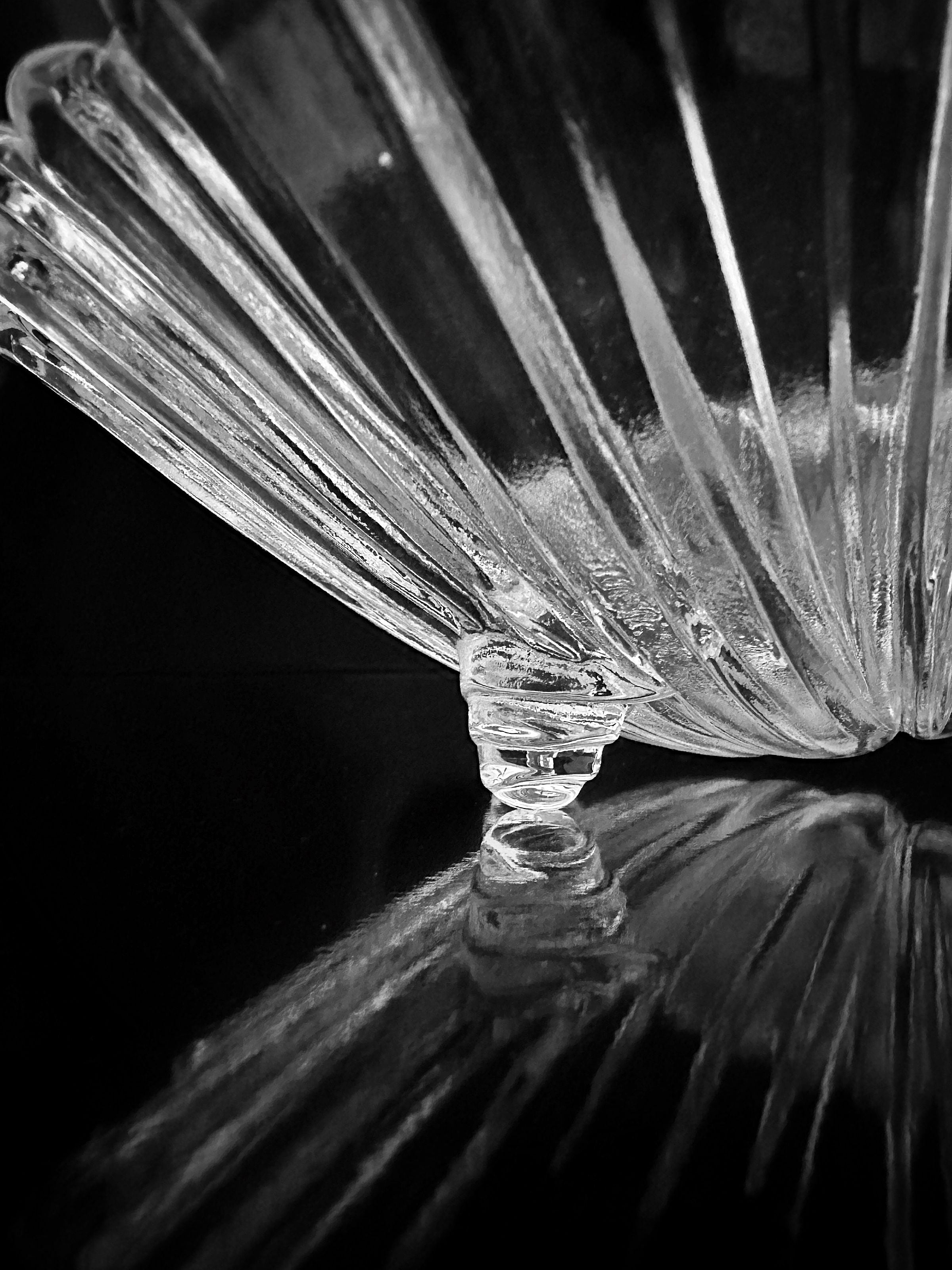 In a black and white image, a 1980s Shell Shaped Glass Bowl by Dodo Vintage is seen resting upside down, its fluted design casting intricate reflections on a dark, glossy surface—ideal for presenting delicacies.