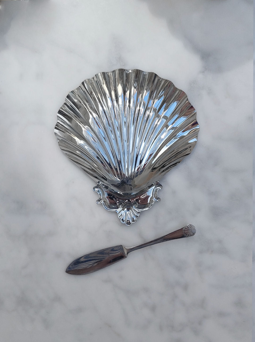 The Shell Butter Dish by Les Objoies features a silver shell design with intricate detailing at the base, and comes with an assorted knife that has a decorative handle, all beautifully showcased on a white marble surface.