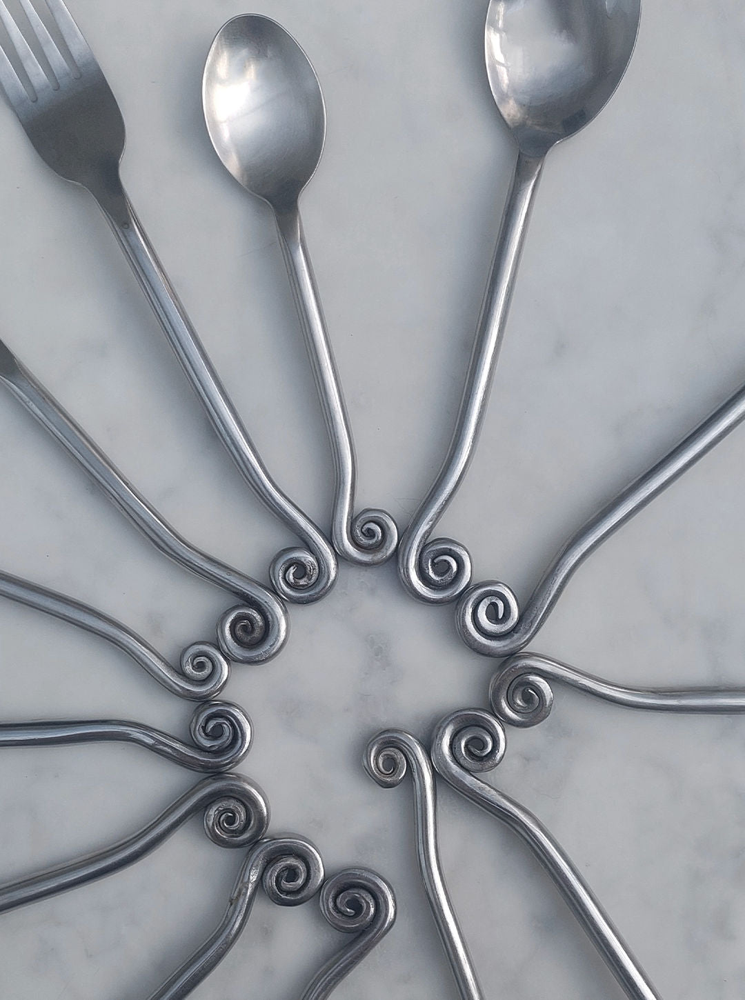 A Set of 4 Swirling Cutlery by Les Objoies, featuring metal forks and spoons with decorative spiral handles, is elegantly arranged in a circular pattern on a marble surface.