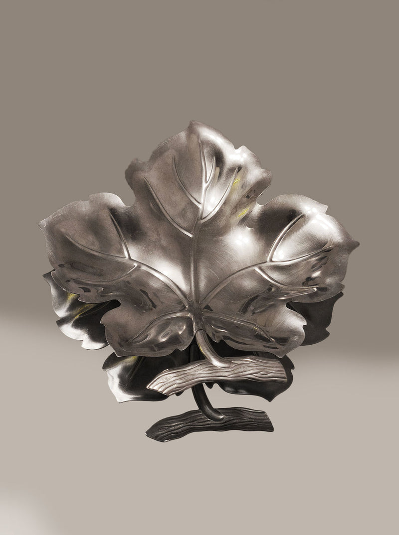 The Wine Leaf Vide-Poche by Les Objoies is a stunning stainless steel sculpture with detailed veining resembling a large maple leaf. Its metallic surface beautifully reflects light, emphasizing its minimalist design as it stands elegantly against a neutral background.