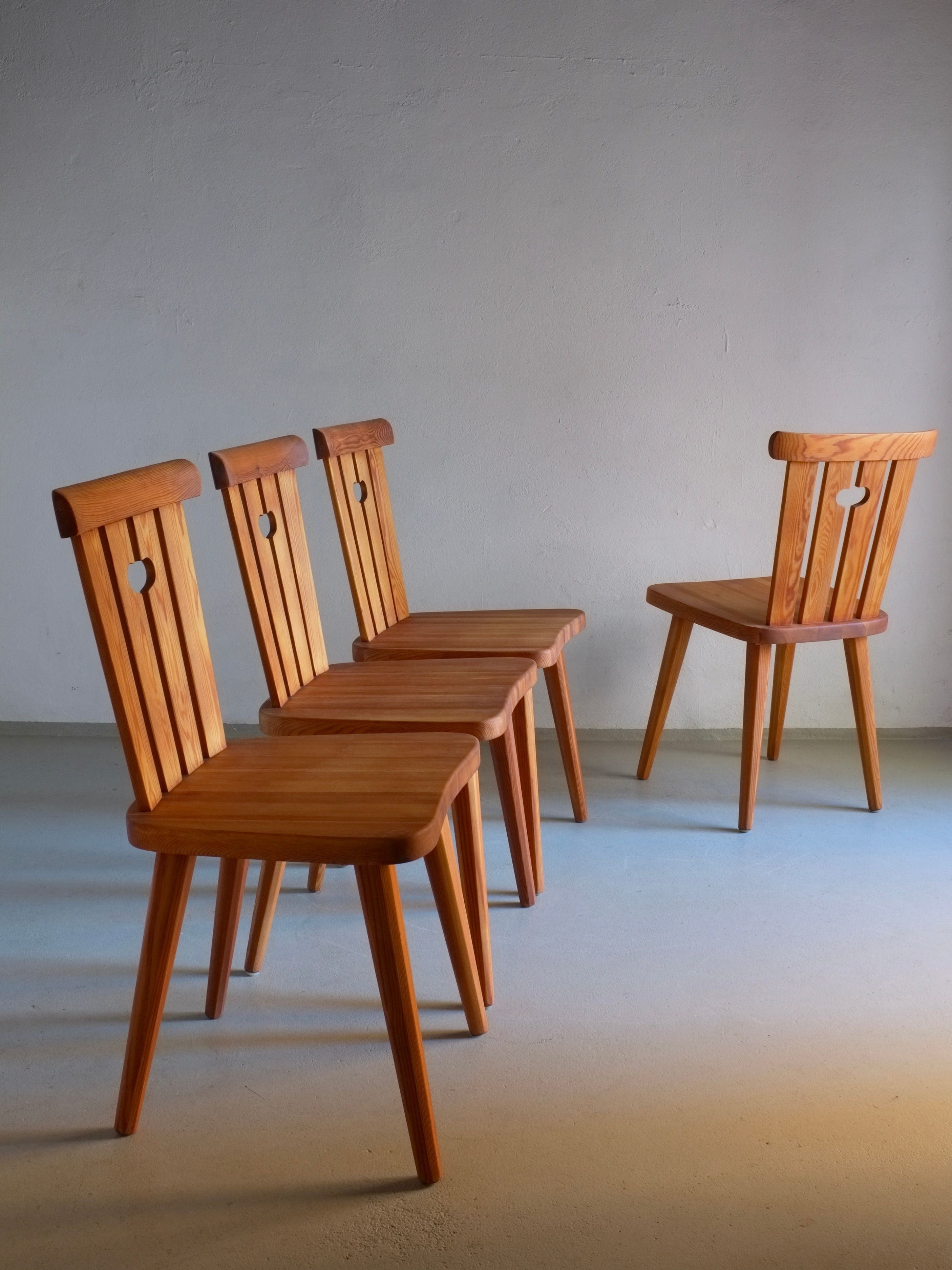 A set of four Rustic Pine Chairs by Goran Malmvall from the 1940s, offered by Veter Vintage and characterized by their heart-shaped cutout designs in the backrests, are arranged in a row with one chair slightly set apart. Each expertly restored and oiled piece enhances the simple, empty room's smooth walls and floors, radiating a minimalist charm.