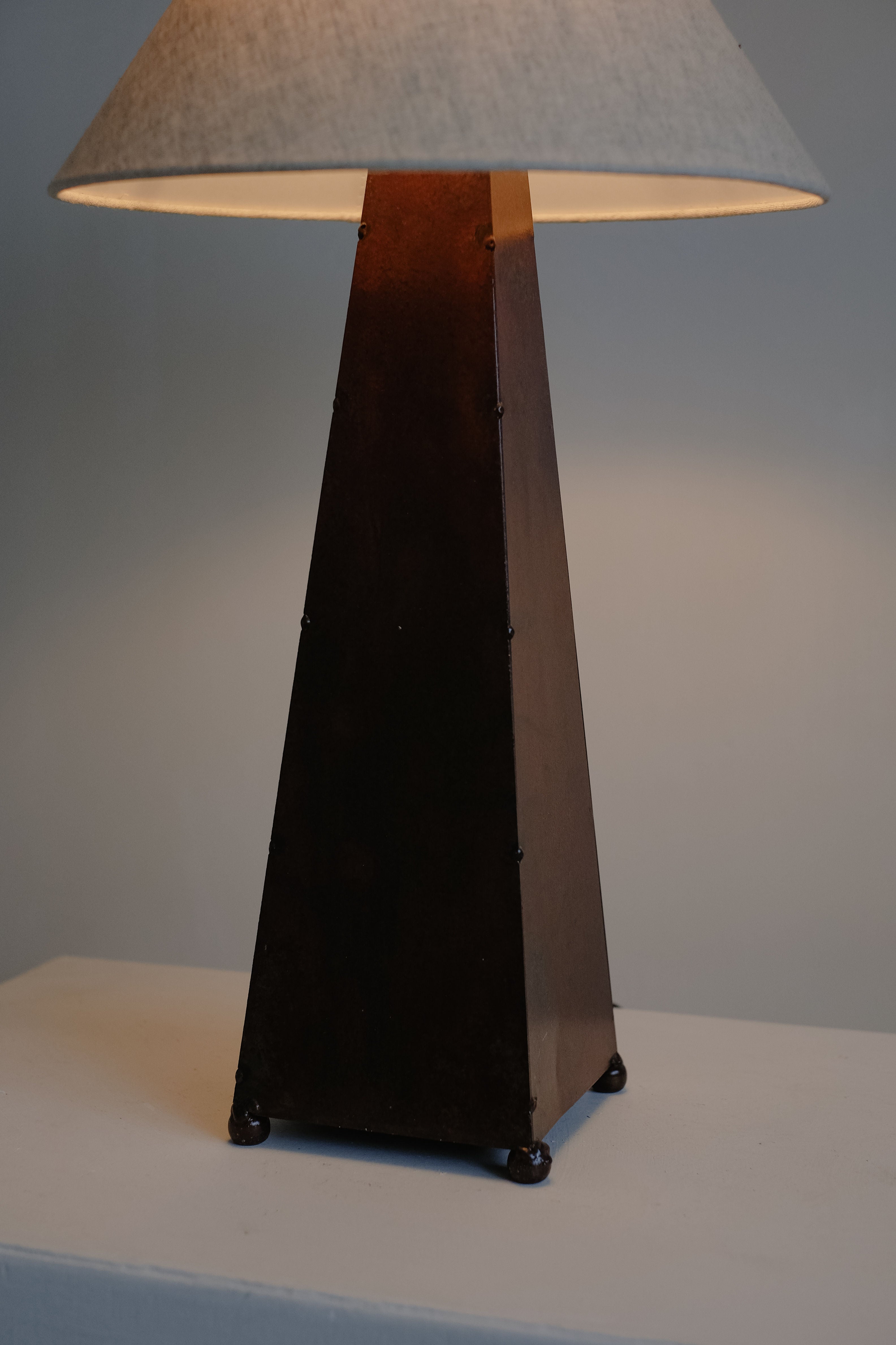 The Obelisk Table Lamp by septembre studios boasts a dark, pyramid shape with a rust-copper patina and beige lampshade, casting warm light. It features a dimmable switch to illuminate any plain white surface against a muted gray backdrop.