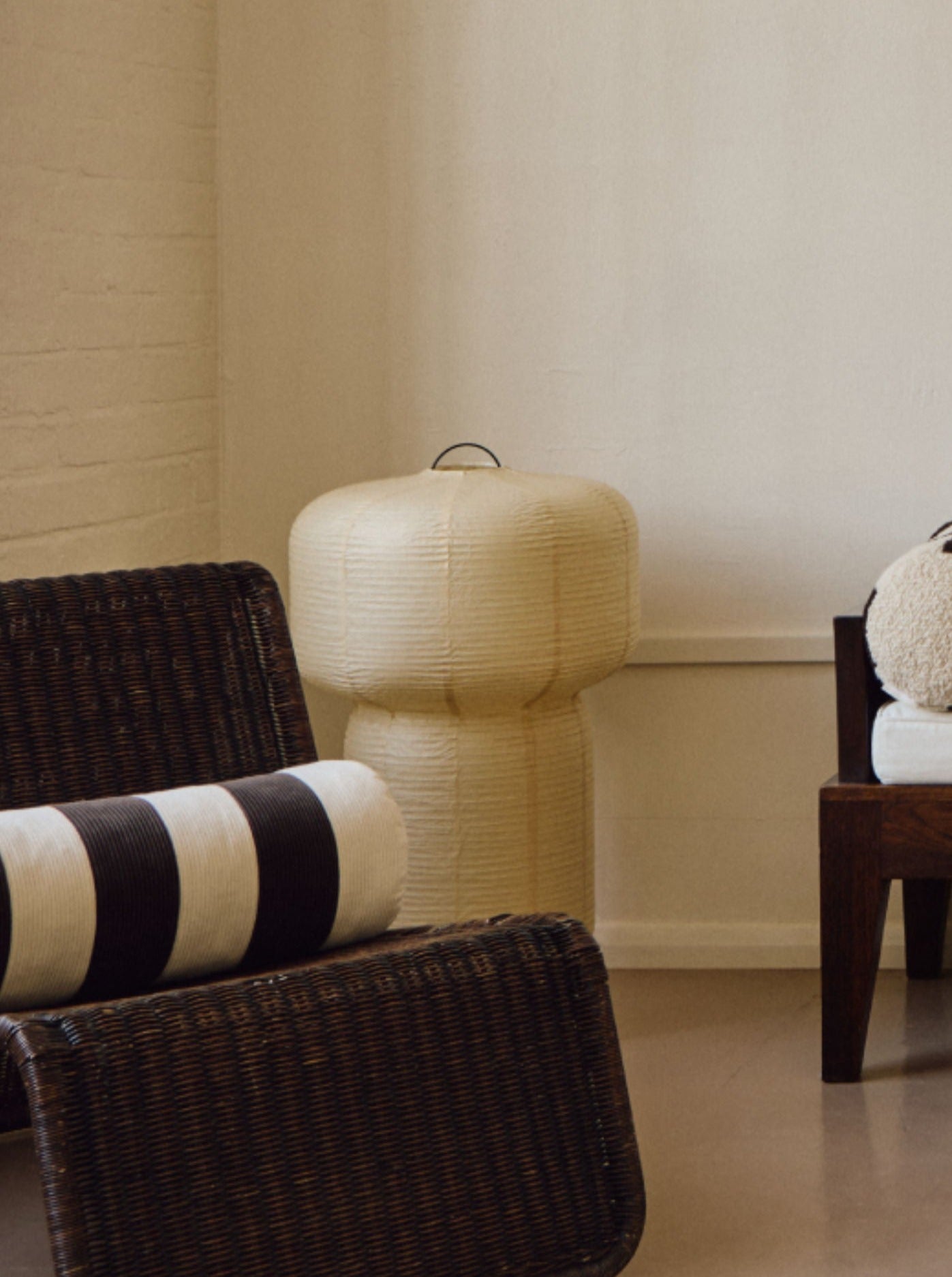 A minimalist room features a wicker chair with a striped cushion, a wooden bench adorned with assorted round and cylindrical pillows in neutral tones, including AMÉLIORÉ's Le Cylindre - Cotton Corduroy - Espresso/Chalk. A textured lamp stands to the side, all against soft cream walls that enhance the warm, tranquil ambiance.