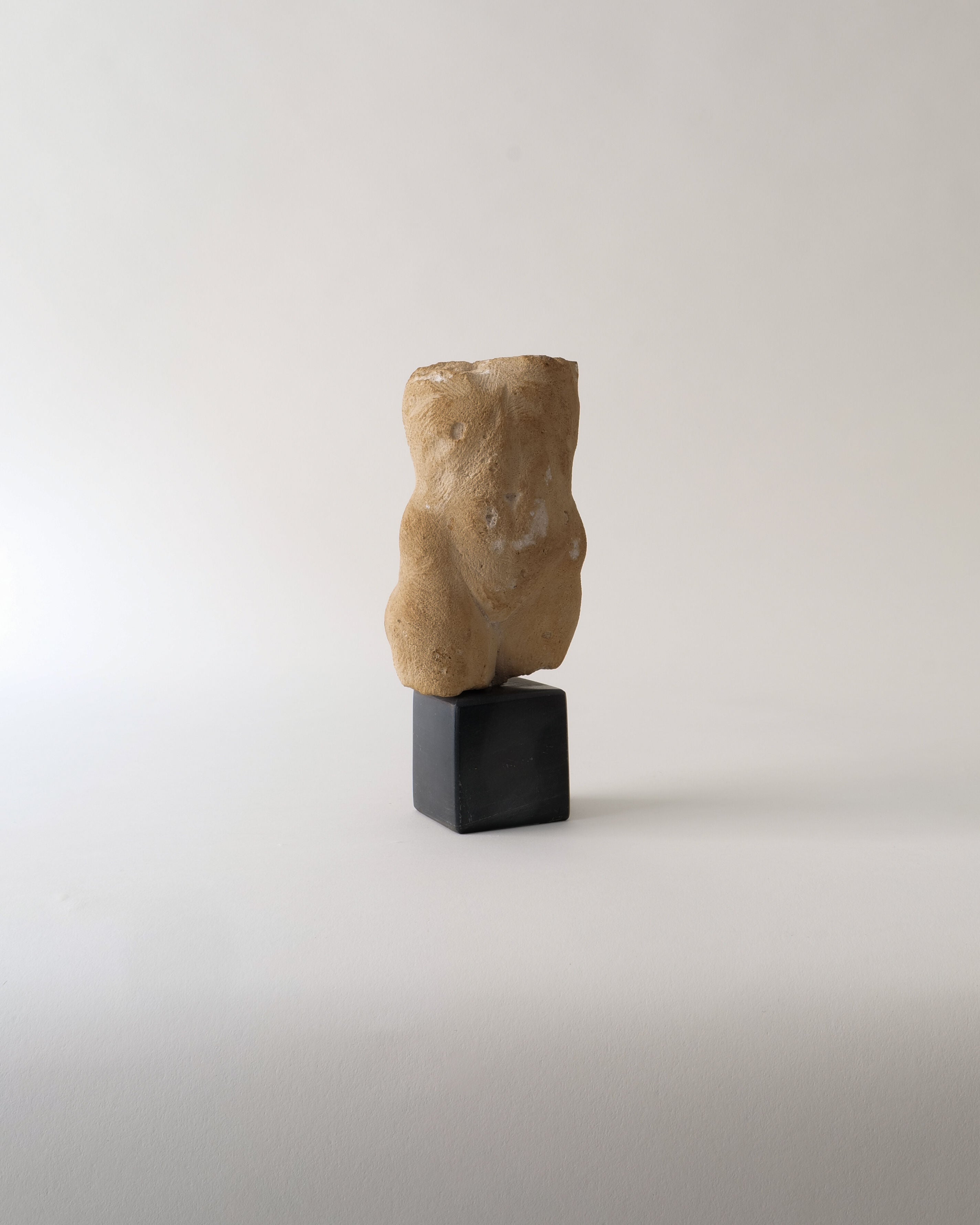 The Torso Sculpture by Misja Kristoffer Rasmussen (b.1971) from Collection apart is a small, abstract beige stone piece resembling a torso, set upright on a black rectangular base against a light, plain background.