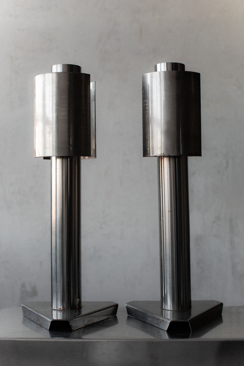 Two tall, cylindrical metal Out For Lunch 1960's Space Age Lamps with a modern design stand on a reflective surface. Each lamp has a vertical base supporting a wider top, creating an elegant contrast against the plain, light-colored wall.