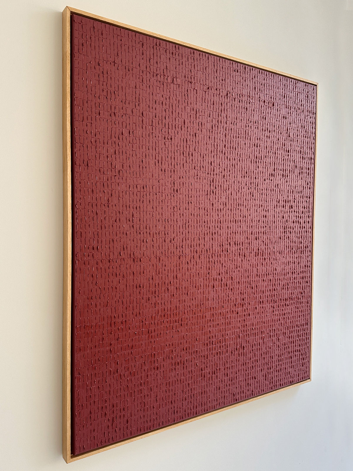 A framed abstract painting hangs on a wall. The artwork, evocative of the Dansaekhwa movement, features a deep red surface with an intricate, slightly raised texture that appears to consist of small vertical and horizontal markings, creating a subtle pattern. The frame is simple and wooden. This piece is Indian Red - Oil on canvas by Roi Elmaliah.