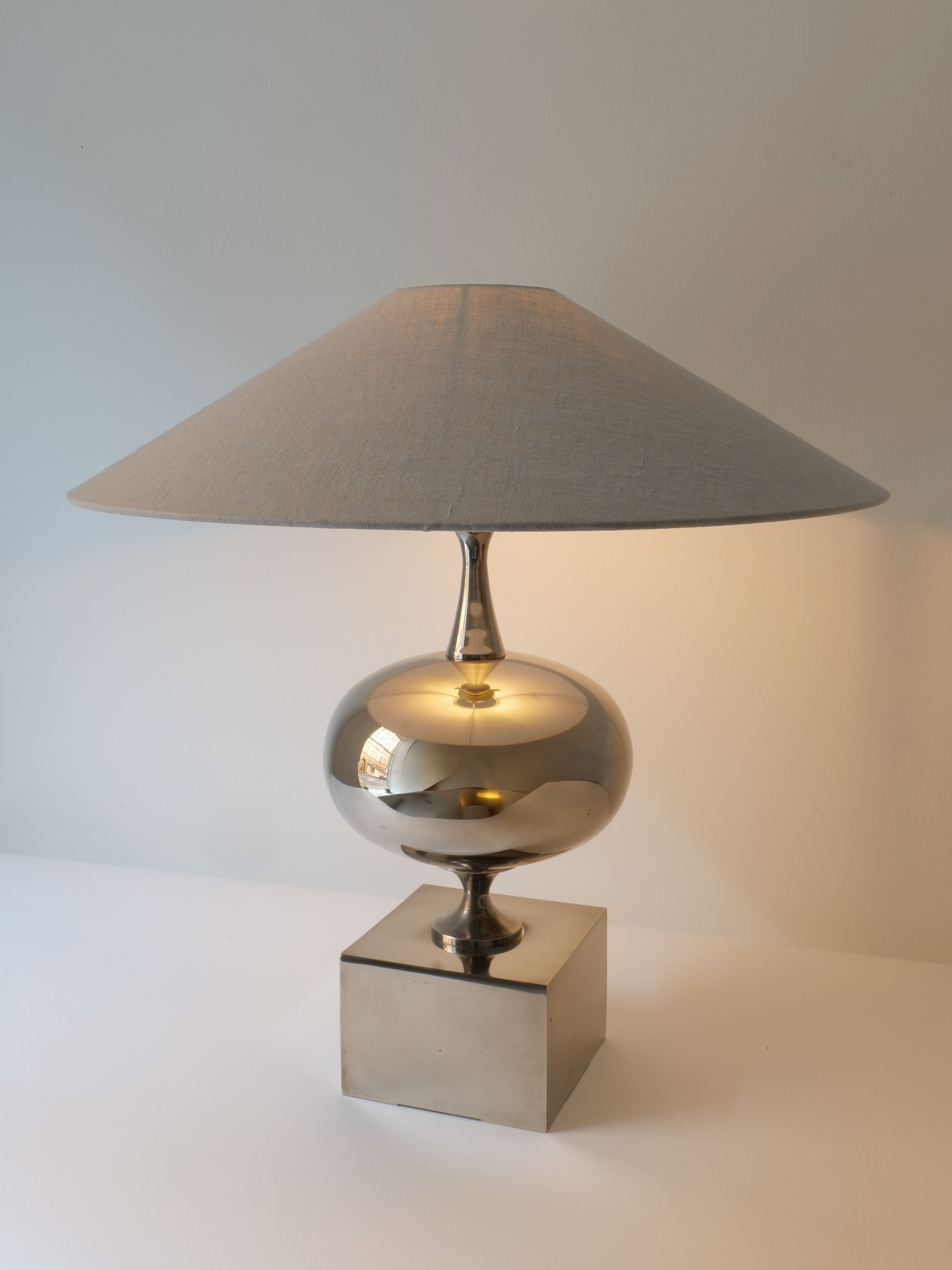 The Barbier Sculptural Lamp 1970s by Collection apart, featuring a metallic, reflective base and a wide, beige fabric shade, is placed on a white surface against a light-colored wall. Reminiscent of 1970s French lighting, the chrome over brass lamp is turned on, emitting a soft, warm glow.