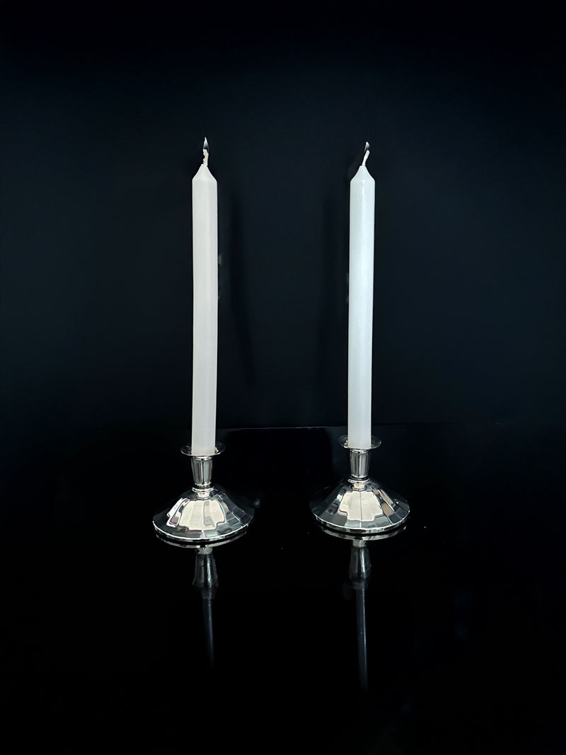 Two tall white candles are positioned in Silver-Plated Art Deco Candleholders from 1930 by Dodo Vintage, standing side by side against a dark backdrop. The candleholders and candles are beautifully reflected on the glossy surface below.