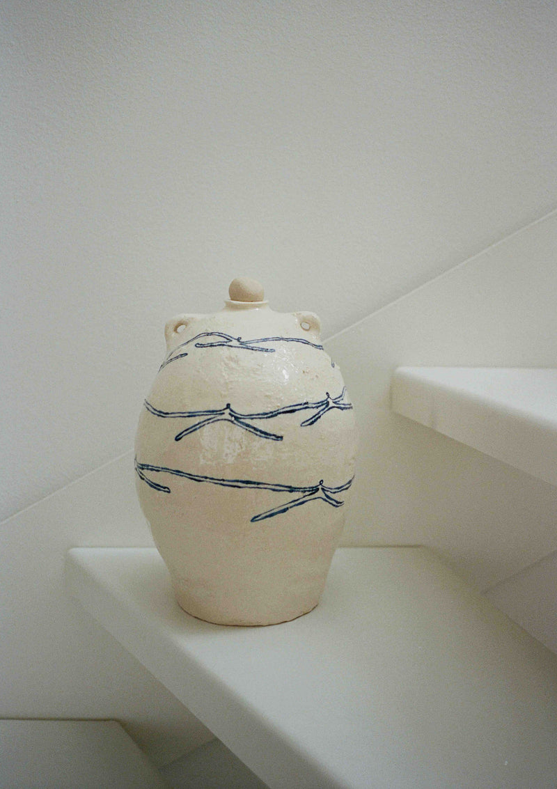 The ‘Ladies Holding Hands’ Ceramic Vase by ATELIER1991, cream-colored with blue dragonfly motifs, sits on a white stair step against a plain white wall.