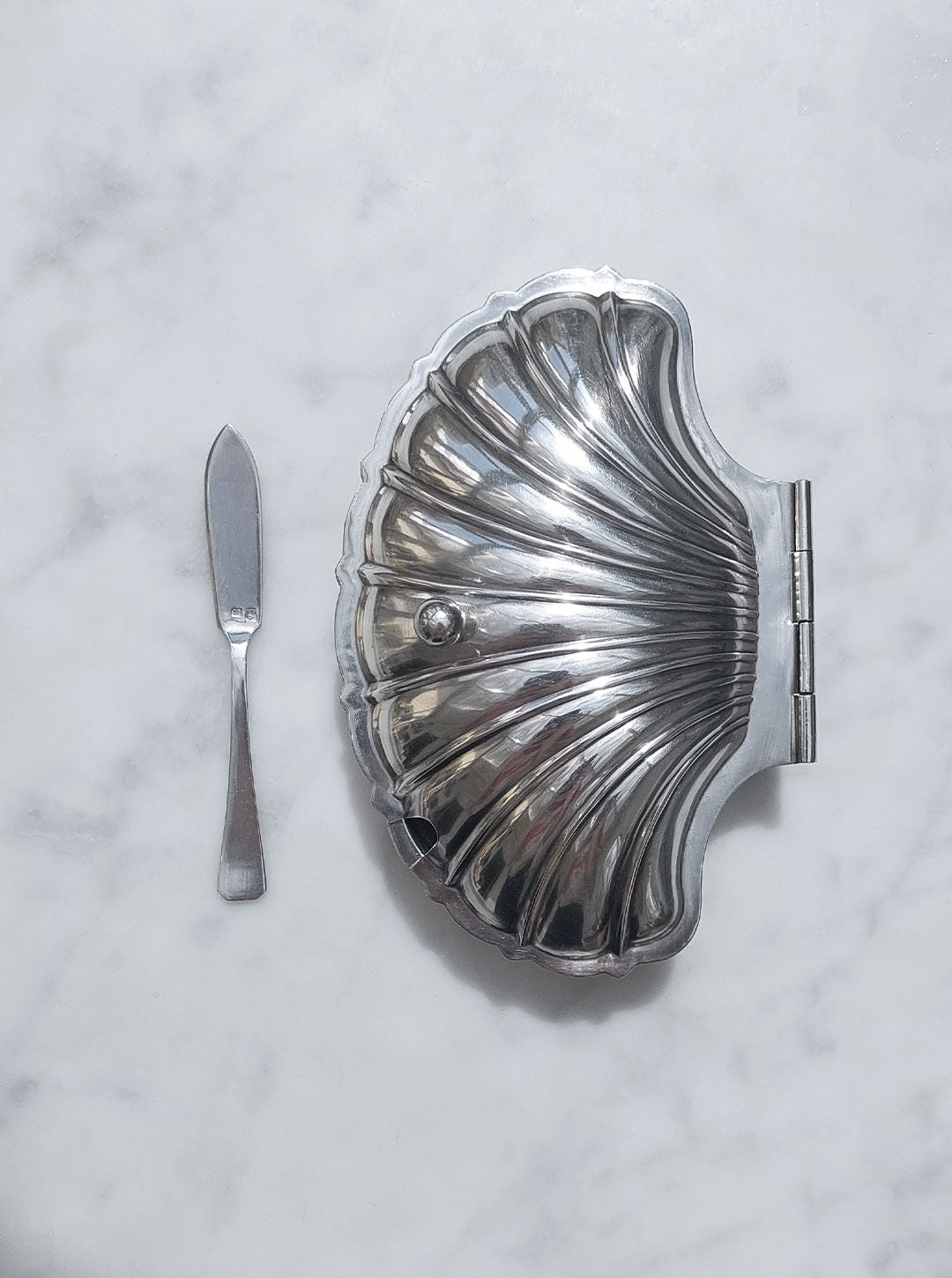 A silver butter knife lies next to a closed, Shell Silver-Plated Butter Dish by Les Objoies with a hinged lid on a marbled white surface. The dish features a polished, reflective finish and a small round handle, exuding vintage charm.