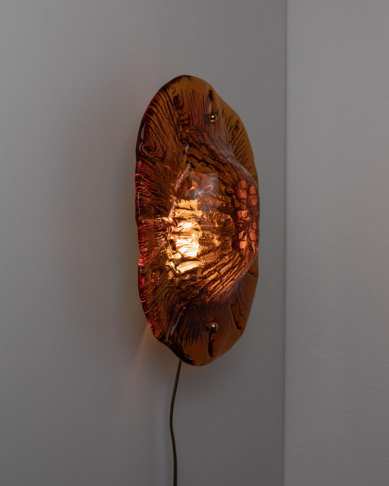 A wall-mounted Vitrika 60s Wall Lamp by Bottega Jacobs, featuring an amber hue and a vintage glass design with a visible bulb inside. This sconce casts a warm glow, accentuating its organic, wavy patterns and adding a nostalgic charm. It is affixed to a plain, light-colored wall.