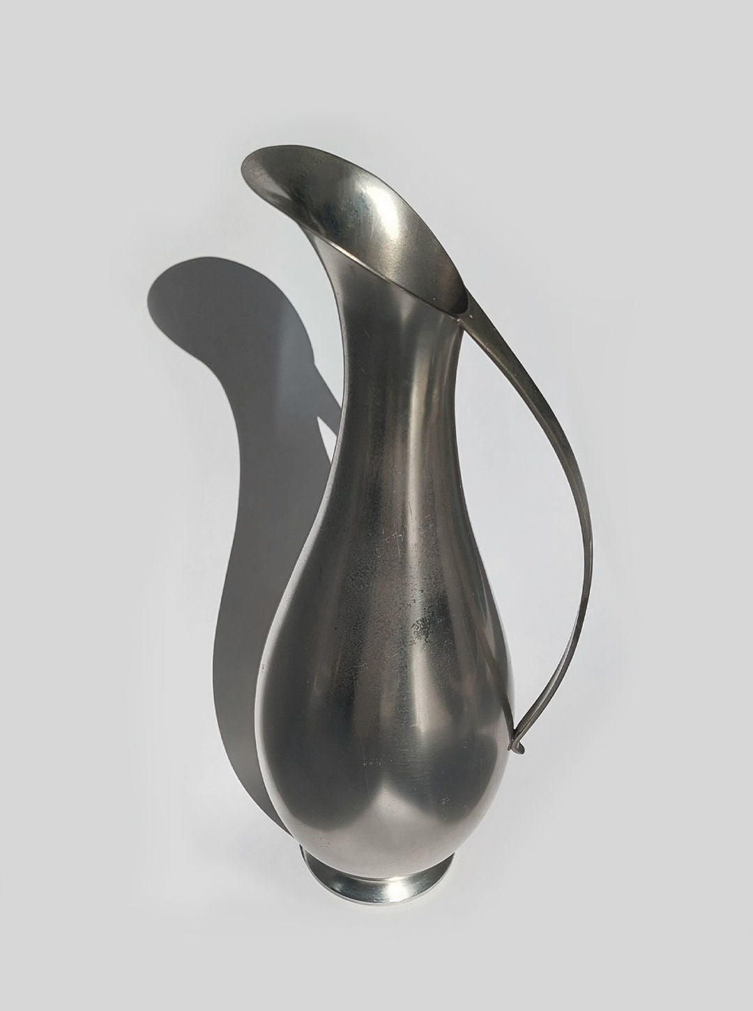 The Large Pewter Carafe by Les Objoies features a minimalist aesthetic with its curved, elongated spout and narrow base, casting a distinct shadow on the plain gray background. Its thin, elegant handle enhances its contemporary design.