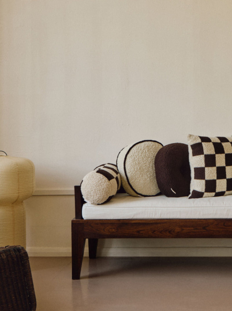 A minimalist room features a wooden-framed daybed against a plain light-colored wall, adorned with handmade cushions in black, white, and brown tones. A wicker lounge chair is in the foreground, and a unique layered lamp stands beside the daybed, adding to the vintage-inspired design. The focal point of the space is topped off with an AMÉLIORÉ Le Rond - Passepoil - Wool Bouclé - Egg White/Chocolate cushion that elevates the overall aesthetic.