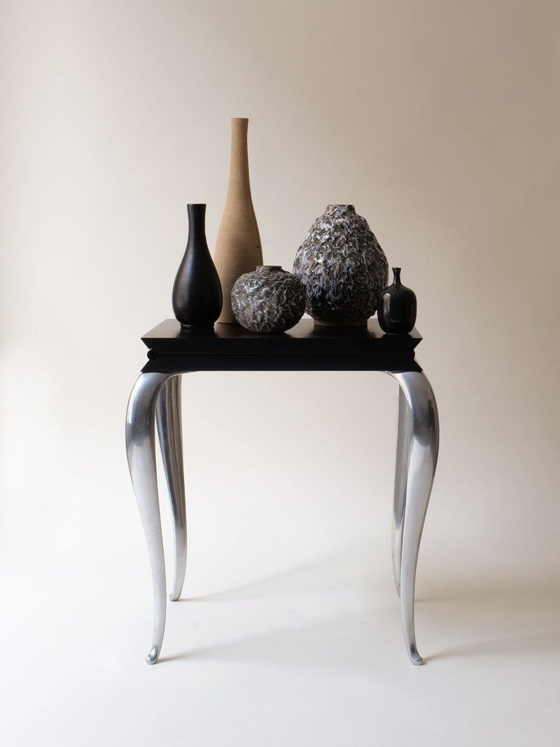 On a modern black table with curved silver legs, five collectable vases of varying shapes in black, gray, and tan sit. Their textured surfaces echo a sleek Louis XV design against a plain cream background. Nearby is the "Lola Mundo Chair" by Philippe Starck (1984) from Collection apart.