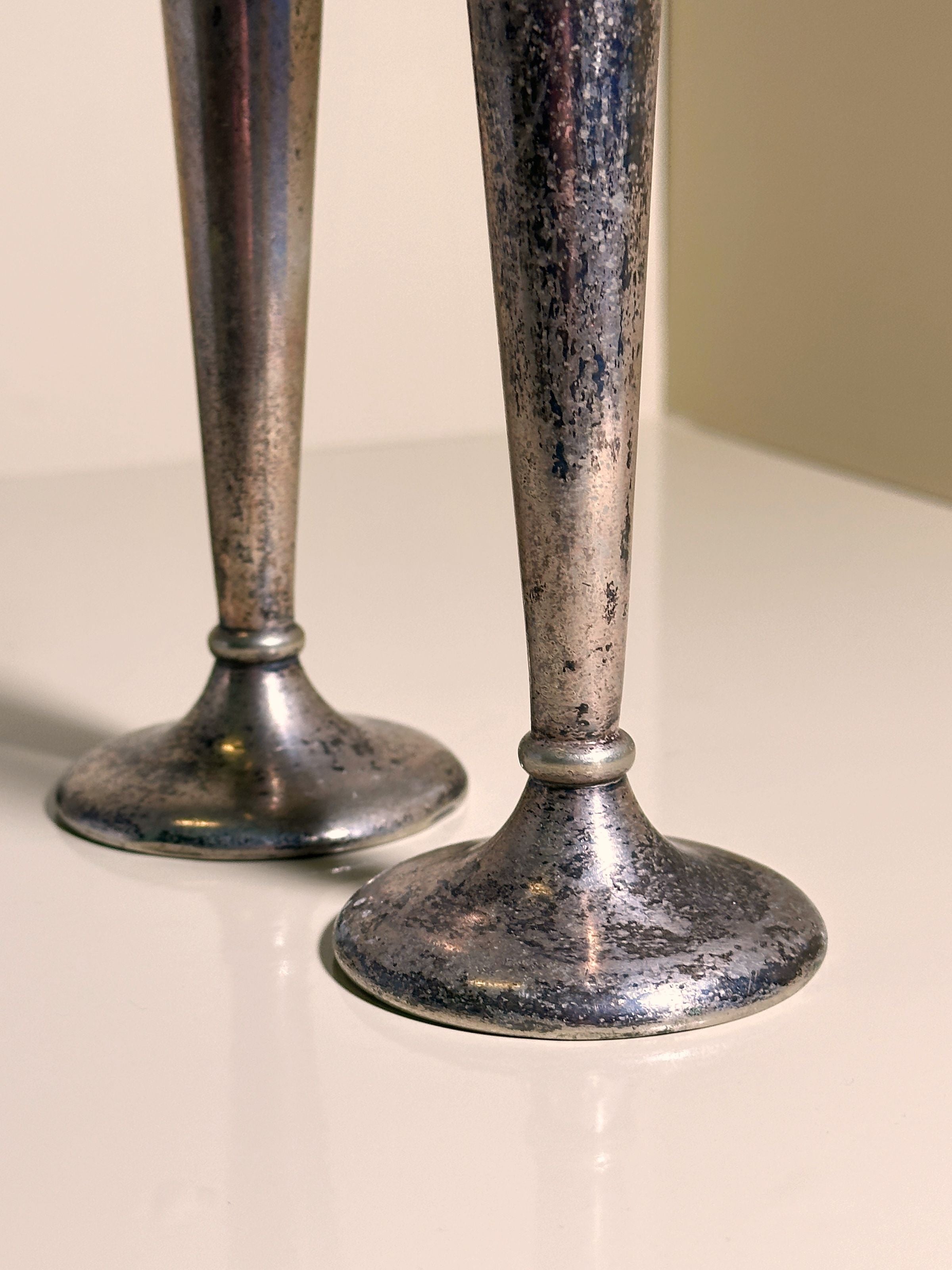 Close-up of the bases of two tarnished Gio Ponti Alpacca silver candlesticks from the 1930s by Dodo Vintage, on a glossy white surface. Soft lighting highlights their textured surfaces as they stand side-by-side casting subtle shadows.