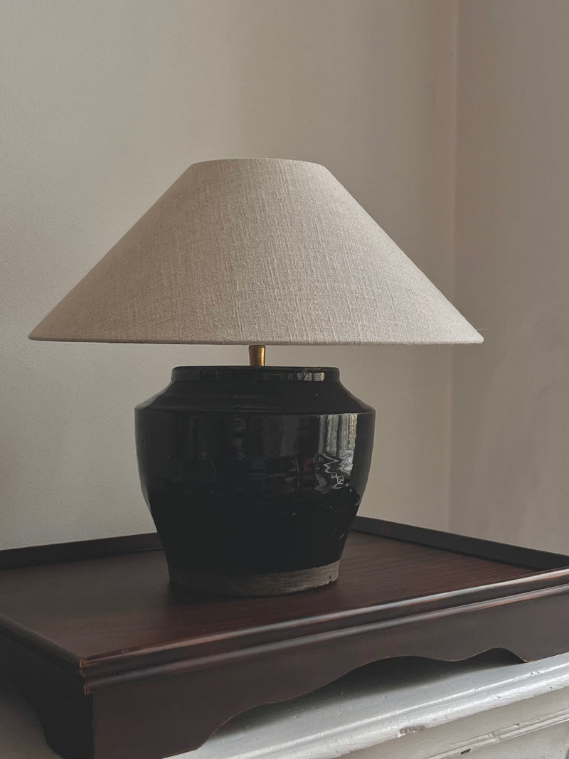 The Médecine Antique Glazed Lamp has a wide beige fabric shade and an antique vessel design with a glossy finish. It sits on a wooden table against a plain wall, seamlessly integrating with its sleek black textile cord for an elegant touch.