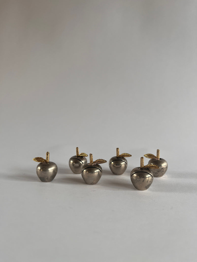 Six Christofle name card holders by Maud Vaughan, metallic apple-shaped with gold-toned stems and leaves, sit elegantly on a smooth light gray surface, contrasting beautifully against the plain light-colored wall.
