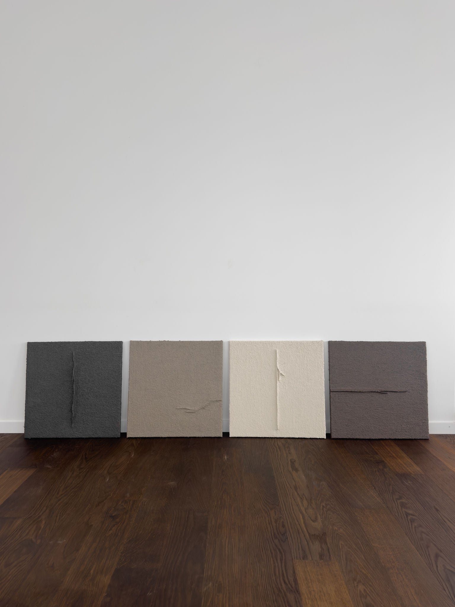 Shadow World 04 by Delal Salazar: Four textured square canvases in dark gray, taupe, off-white, and brown with subtle raised creases, crafted with acrylic and sand on canvas, lean against a white wall on a dark wood floor.