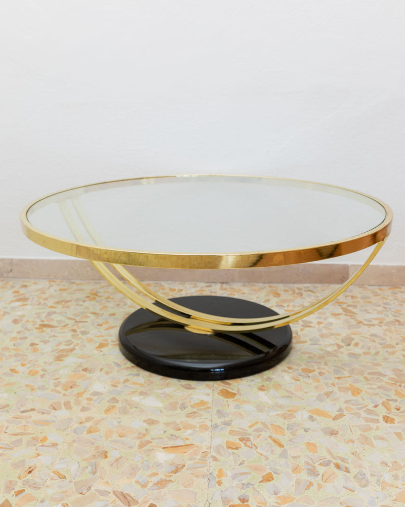 The Glass and Metal Side Table - Italy 1980s by Bottega Jacobs features a round glass tabletop elegantly supported by a gold metal frame with an abstract, geometric base resting on a black circular platform. Its Hollywood Regency style provides a striking contrast against the terrazzo floor and plain white wall.