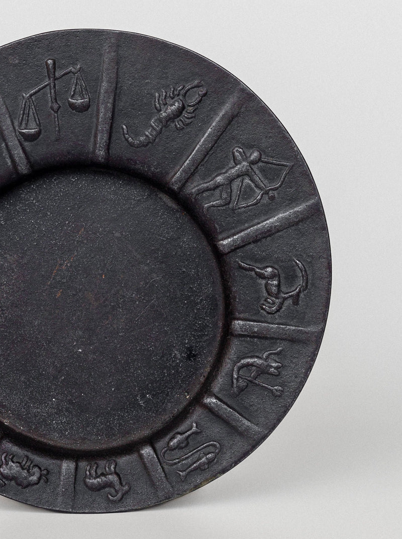 The Art Deco Zodiac Signs Decorative Dish by Spigel is a dark circular plate with vintage iron dish features, showcasing relief carvings of astrological symbols along its rim. The visible symbols include Libra (scale), Scorpio (scorpion), Sagittarius (archer), and Capricorn (sea-goat). This piece embodies Scandinavian vintage design with a plain center.