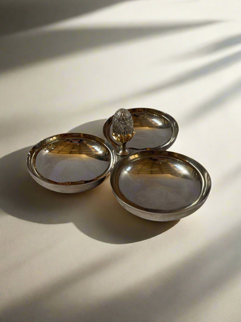 A polished silver trinket dish with three circular compartments connected together. At the intersection of the trays, there is a decorative finial shaped like a small pineapple. The surface reflects the light, highlighting the smooth metal finish of this charming Maud Vaughan Vintage French Nickel and Brass Serving Dish.