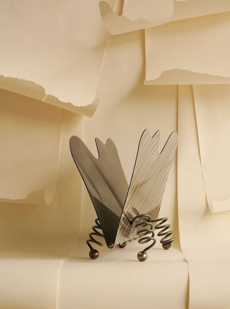 Abstract art installation featuring two curved, dense metal panels resembling butterfly wings, set against a beige backdrop with torn paper layers showcasing the Les Objets Amsterdam Wavy magazine rack.