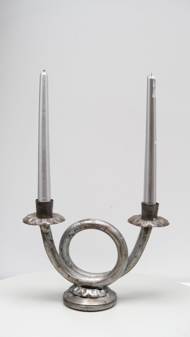 A silver Double Wrought Iron Candlestick with two thin, unlit taper candles. The candlestick, by LA NOUVELLE GALERIE, features an ornate circular design at the base with two arms extending upwards on either side, each supporting a candle. Its dimensions stand out against the plain white background.
