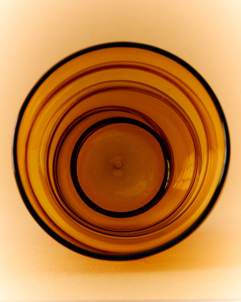 A top-down view of the 'Paguro' Amber Glass by Ylaria Pavone on a light beige surface, showcasing its handcrafted concentric patterns inspired by Italian glassblowing.