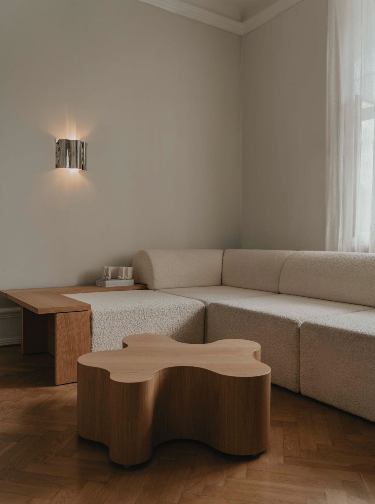 A minimalist living room with a beige curved sofa, a unique wooden coffee table with an organic shape, and a handmade wall light emitting soft illumination from the Psychedelic Wall Scone by Caia Leifsdotter. The side table holds a tea set, the parquet flooring shines subtly, and a sheer white curtain partially covers the window.