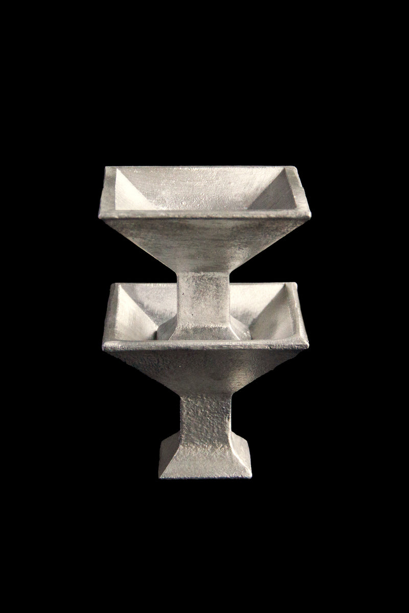 The 'Kare' Serving Coupe by Atelier Barb is a symmetrical, metallic piece made from upcycled aluminum with two stacked square funnels and a central column. Its silver-gray, textured finish highlights sustainability against a black backdrop.
