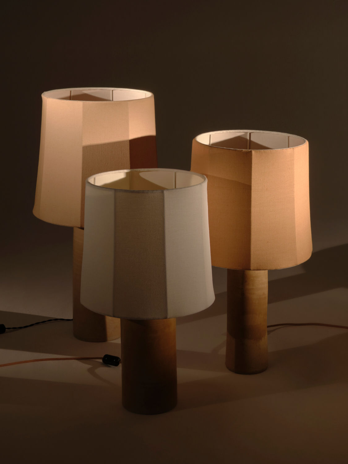 Image of three Nassi Lamps Sonora Linen Table Lamps with beige and white pure linen shades and cylindrical wooden bases, placed closely together in a dimly lit setting. The lamps are turned off, and their power cords and switches are visible on the surface, evoking a warm ambiance reminiscent of Puglia.